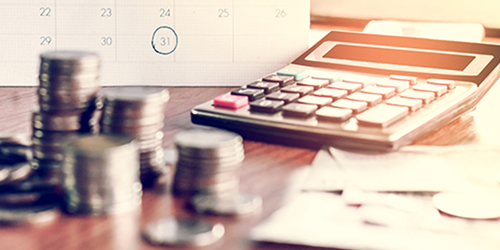 Stacks of coins and a calculator sitting on a desk in front of a calendar with a date circled, symbolizing a payment date.