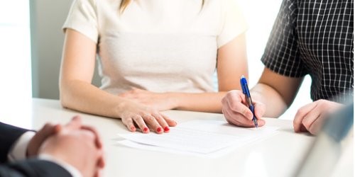 Image shows two torsos: a feminine and a masculine, sitting together at a white table. There are papers on the table and the masculine arm holds a pen positioned as if to sign a document. The feminine arms are resting on the table and red nail polish is featured. In the lower left corner you can see two additional masculine hands clasped together. This is all you can see of the people. 