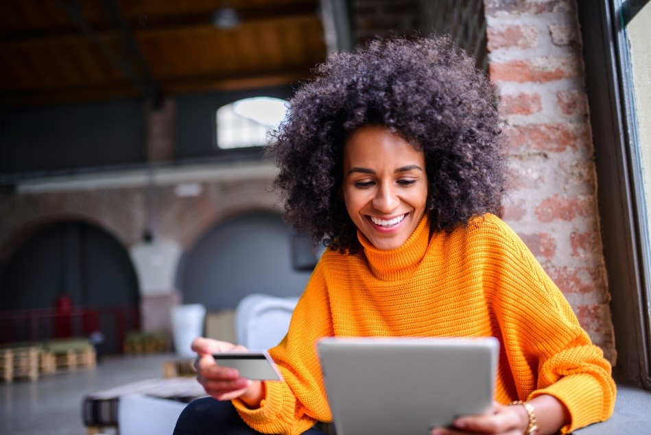Woman in orange turtleneck sweater is sitting in a brick warehouse type space, holding a tablet in her left hand and a credit card in her right. She is smiling and looks like she might be reading the information on the front of her credit card to do some online shopping. 