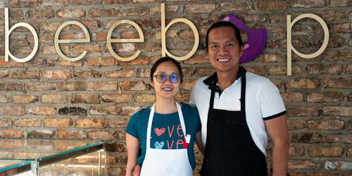 Pauline Nario and Alexander Benedicto, the owners of BeeBop Doughnut Shop in Calgary, both smiling in front of the beebop signage of their shop.