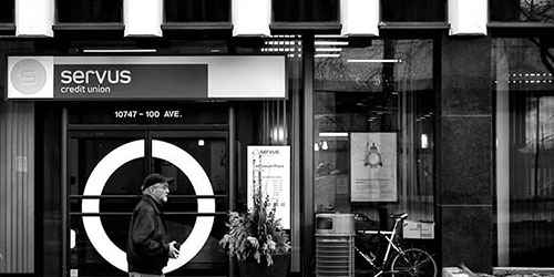 Black and white image of man walking down the street in front of a Servus Credit Union branch. Photo taken by George Stastny.